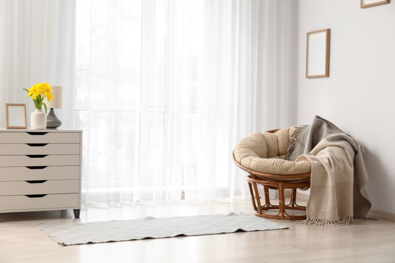 Cortina de voil em porta balcão, decorando a sala de estar e contrastando com as texturas da poltrona em madeira e tecidos ásperos.