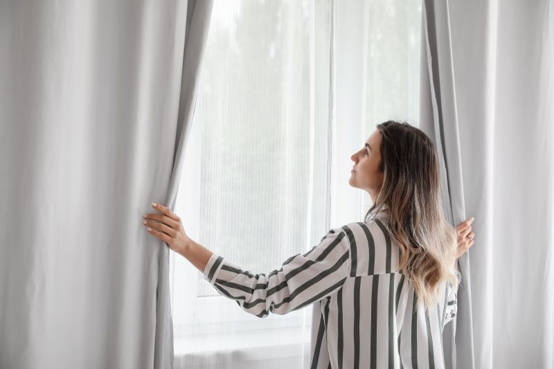 Mulher abrindo a cortina de um cômodo, representando a importância da iluminação na decoração.