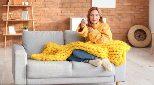 Mulher sentada em um sofá cinza, coberta por uma manta grossa amarela e um casaco amarelo aponta o controle para a televisão.