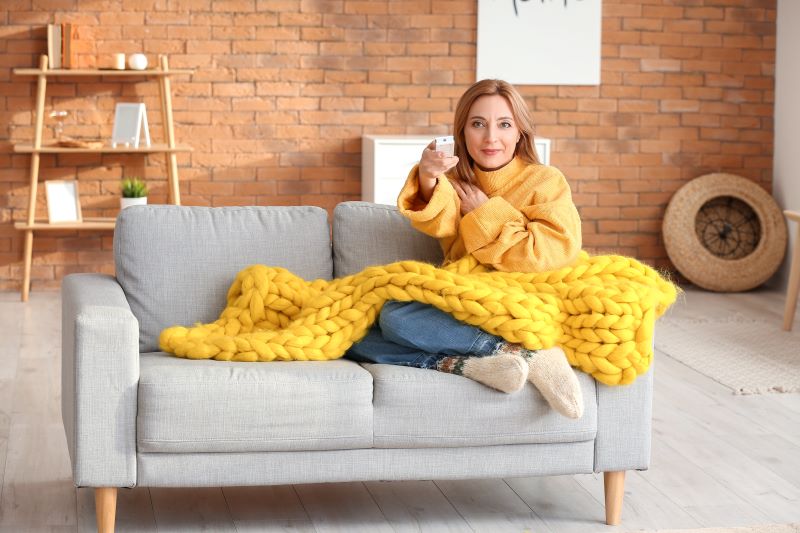Mulher sentada em um sofá cinza, coberta por uma manta grossa amarela e um casaco amarelo aponta o controle para a televisão.